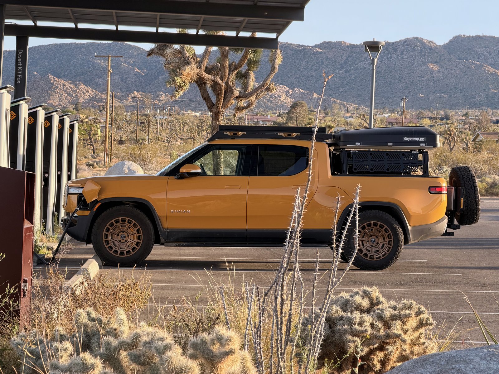 The loaded Rivian charging at sunset, side on, with roof rack, bed rack, rooftop tent, sliders and spare
